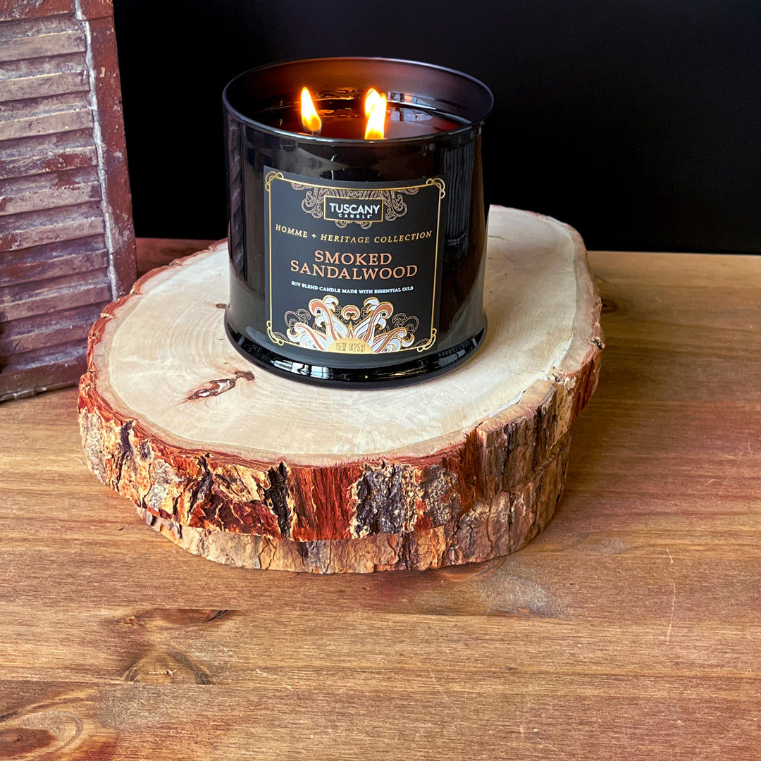 A Smoked Sandalwood scented candle from the Homme + Heritage Collection by Tuscany Candle sits on top of a piece of wood.